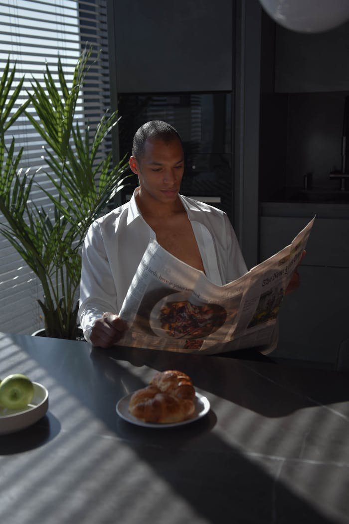 Man in White Unbuttoned Shirt Reading a Newspaper While Sitting in the Kitchen